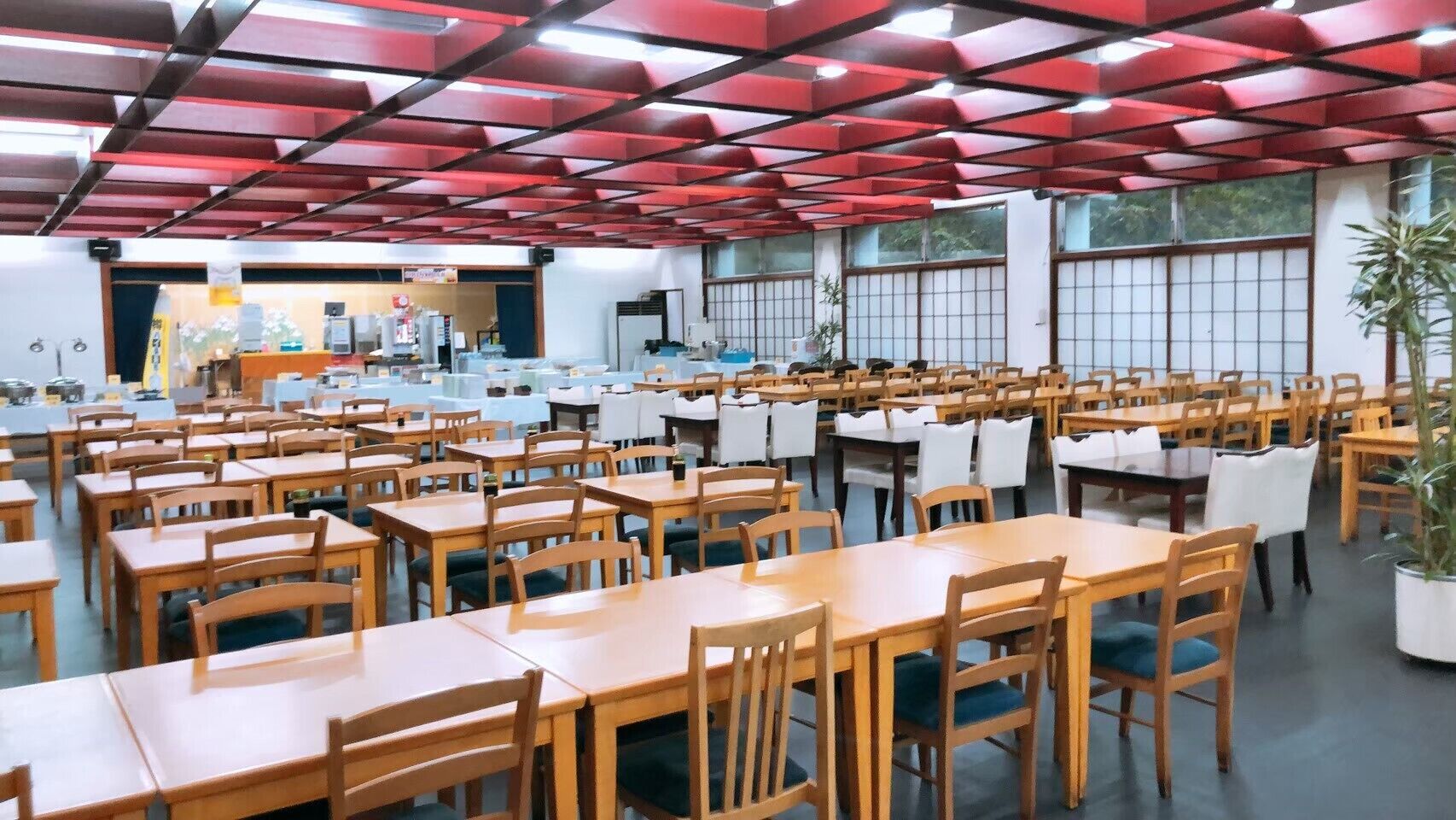Uma sala de jantar espaçosa com várias mesas e cadeiras, teto vermelho e janelas amplas.
