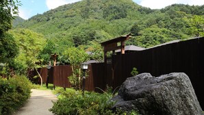 Ein Pfad führt zu einer Holzhütte mit geneigtem Dach, umgeben von üppiger Vegetation und einer Berglandschaft.