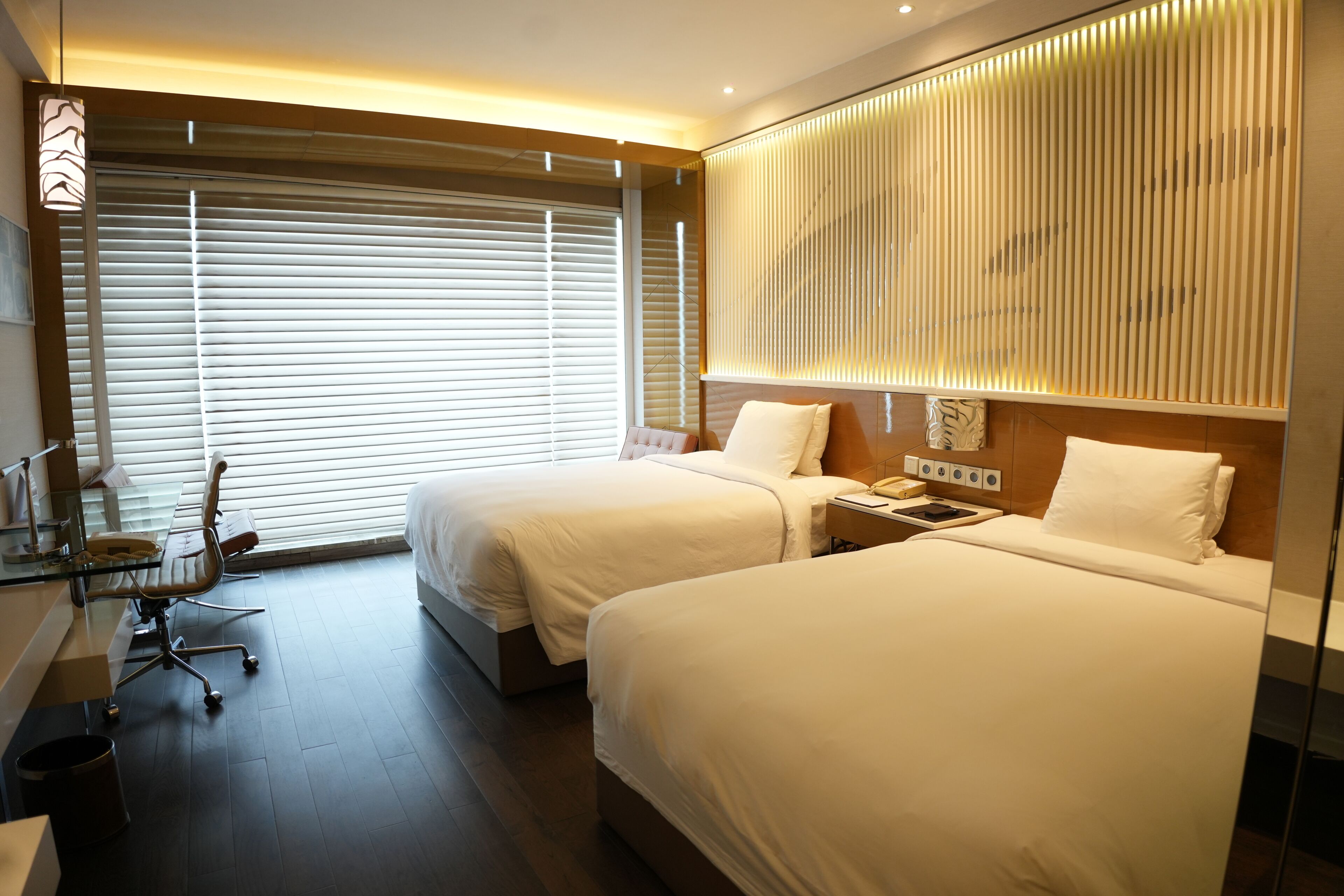 A hotel room with two beds, a desk, and a large window with blinds.
