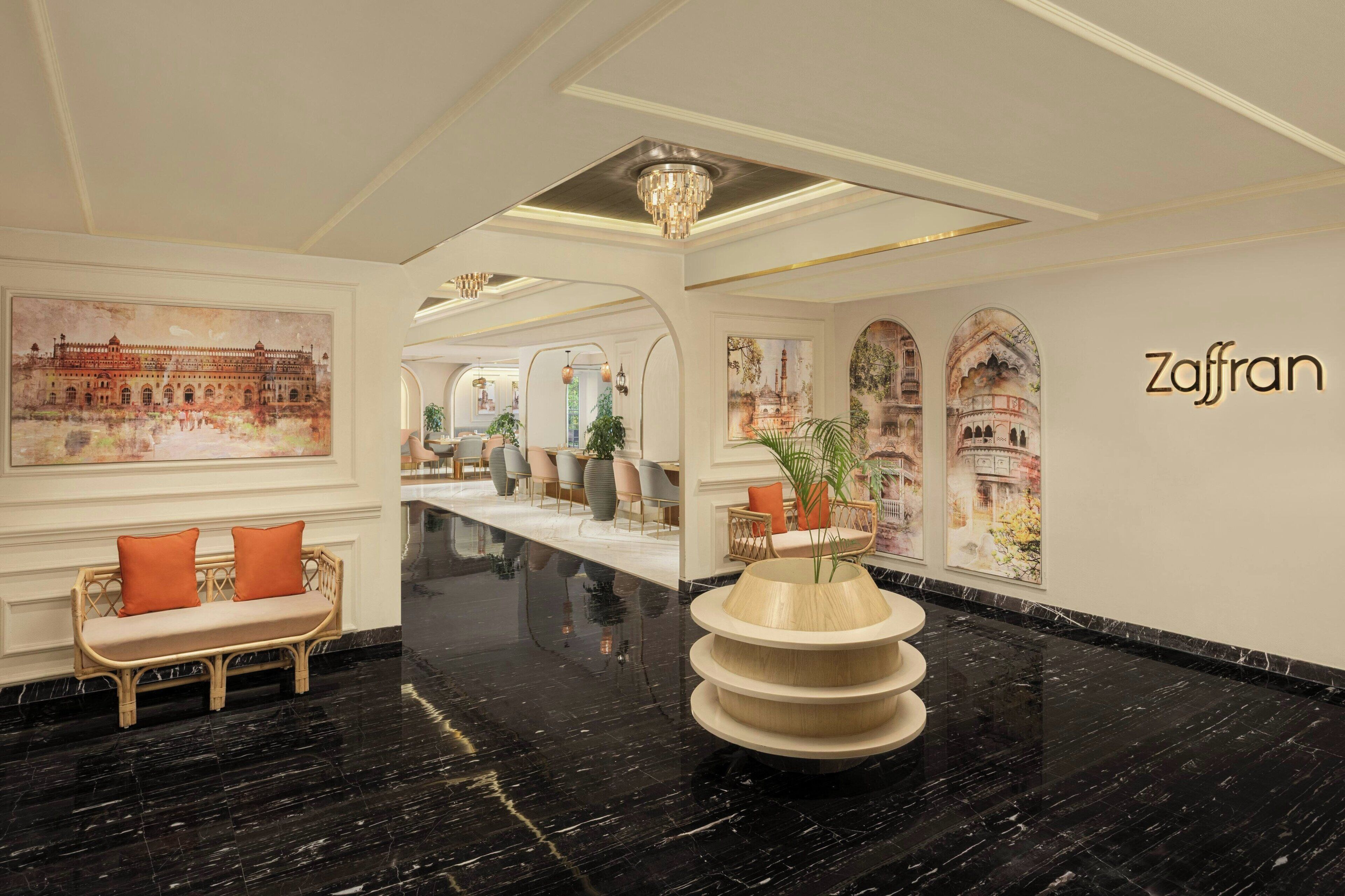 A lobby with a black marble floor, a wicker sofa with orange cushions, a central wooden planter, and wall art depicting buildings.
