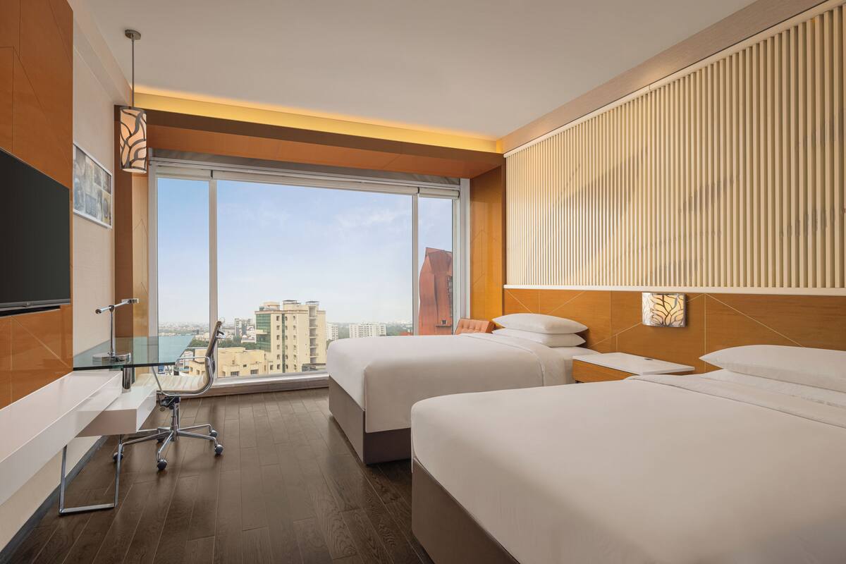 Habitación de hotel con dos camas, un escritorio con silla, un ventanal con vistas a la ciudad y un televisor de pantalla plana montado en la pared.