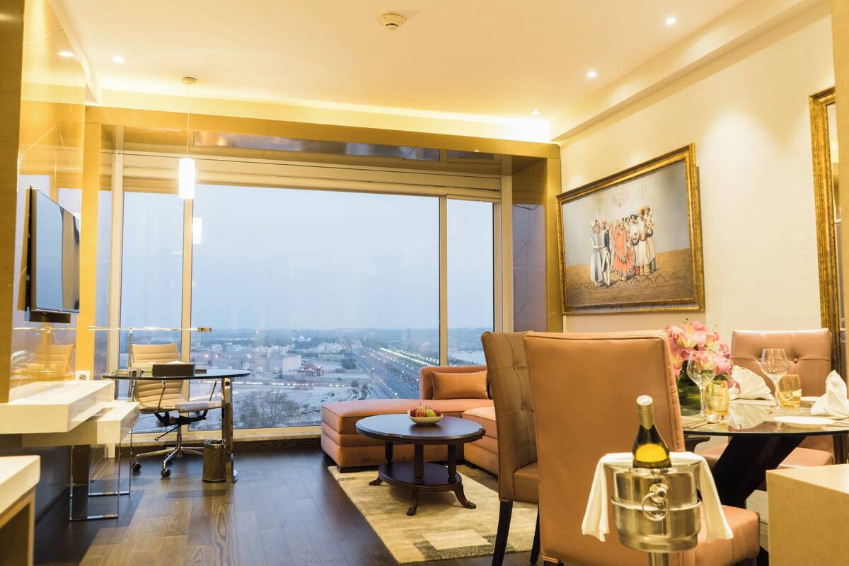 Una habitación de hotel moderna con un ventanal grande, zona de comedor, sofá, televisor y un cuadro en la pared.