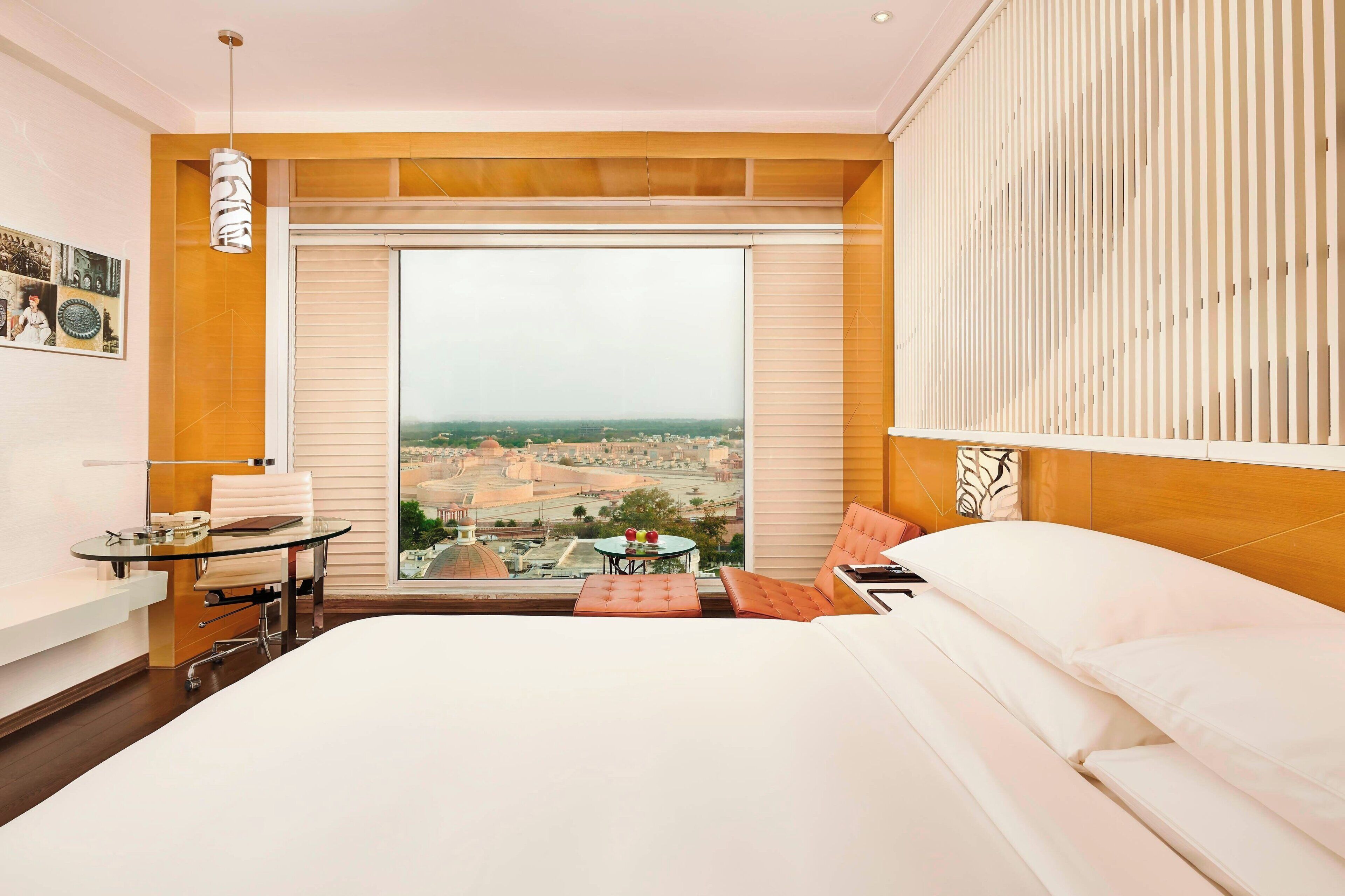 A hotel room with a large bed, a desk with a chair, a small table with a chair, and a view of a cityscape through a large window.