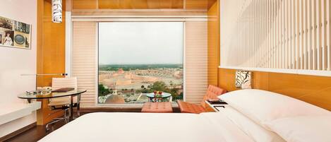 A hotel room with a large bed, a desk with a chair, a small table with a chair, and a view of a cityscape through a large window.