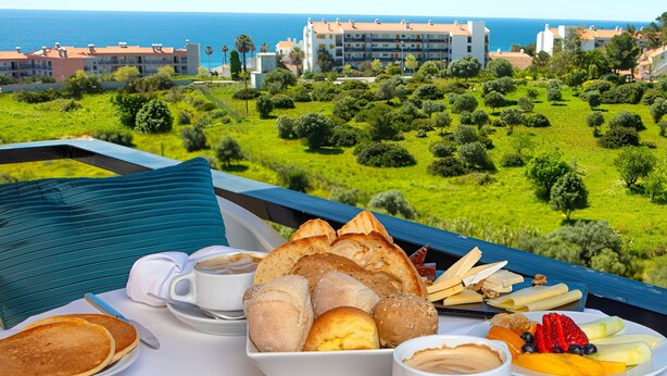 Una mesa de desayuno con pasteles, frutas y embutidos, con vistas a un paisaje costero y edificios.