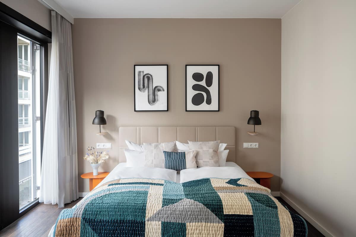 Quarto de hotel com cama, duas obras de arte abstratas emolduradas, mesa de cabeceira com abajur e janela com cortinas.
