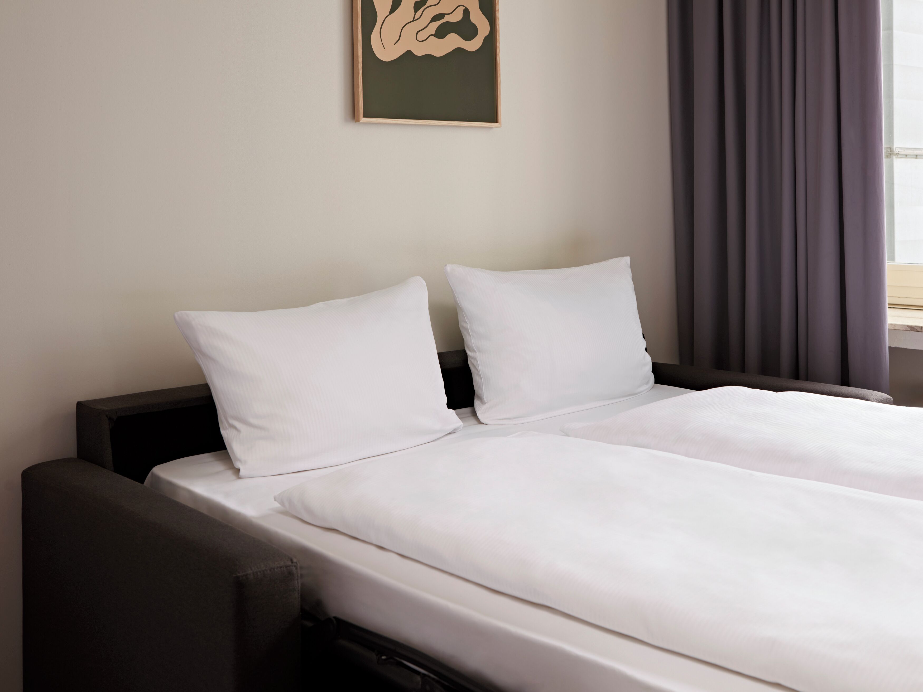 A hotel room with a bed, two pillows, a headboard, a nightstand, and a wall-mounted artwork.