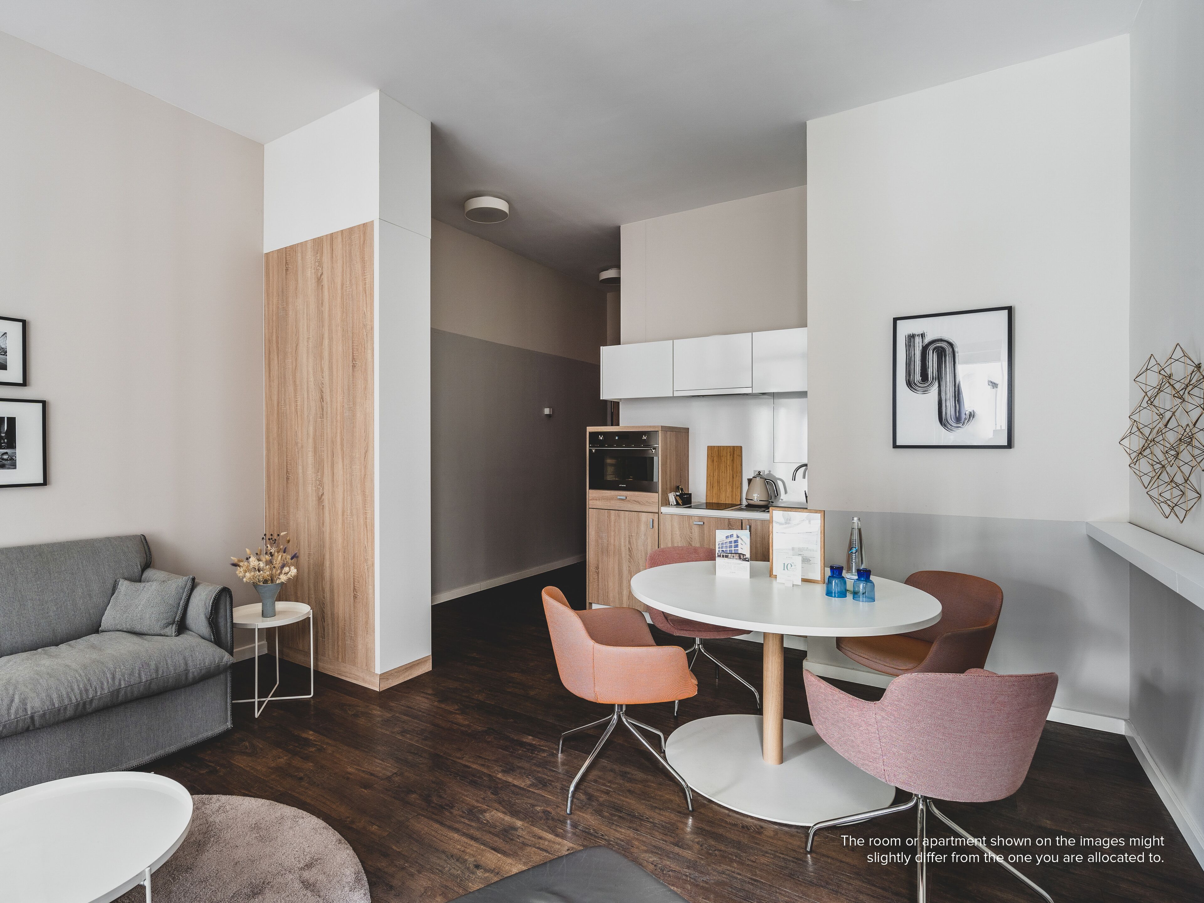 A modern living room with a grey sofa, a round dining table with pink chairs, a small side table with a vase, and a kitchen in the background.