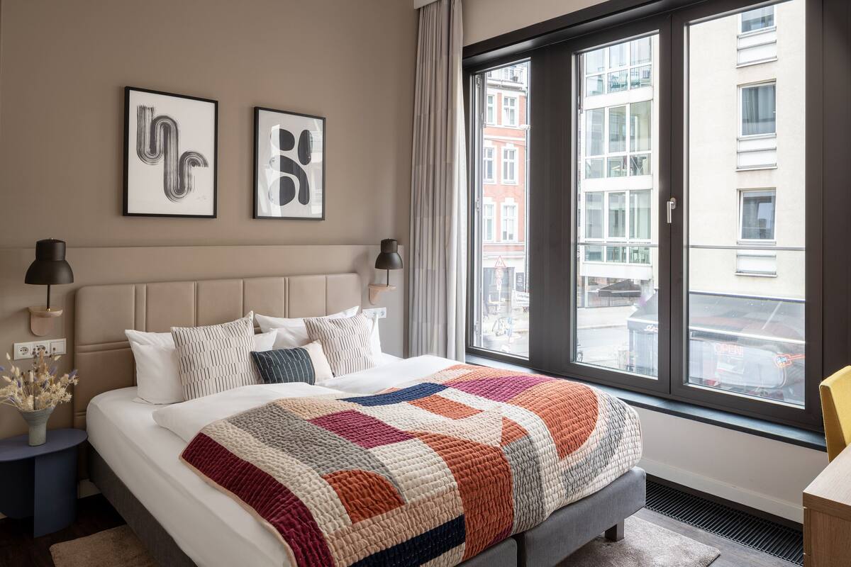Quarto de hotel moderno com uma cama grande, vista da cidade através de uma janela ampla e duas obras de arte abstrata emolduradas na parede.