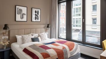 Et moderne hotelværelse med en stor seng, udsigt over byen gennem et stort vindue og to indrammede abstrakte kunstværker på væggen.