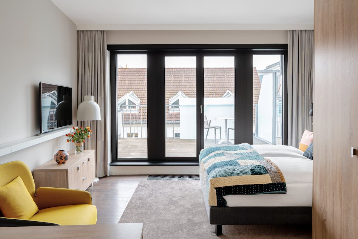 Quarto moderno com uma cama grande, uma poltrona amarela, uma televisão e vista para o exterior.