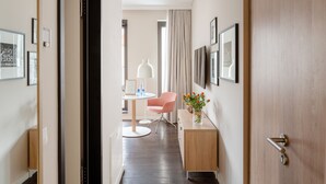 Un corridoio d'hotel con una porta in legno, una scrivania con un vaso di fiori, una sedia rosa e quadri appesi al muro.