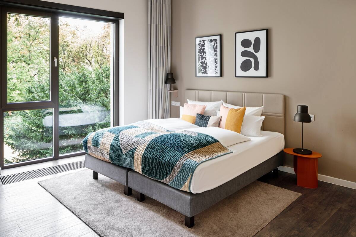 Quarto moderno com uma grande janela, uma cama cinza e vista para árvores.
