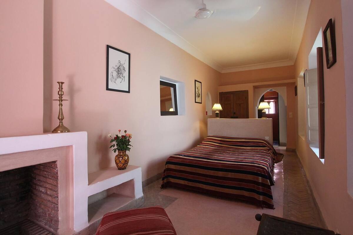 A hotel room with a fireplace, a bed with striped bedding, a bedside table with a vase of flowers, and framed artwork on the walls.