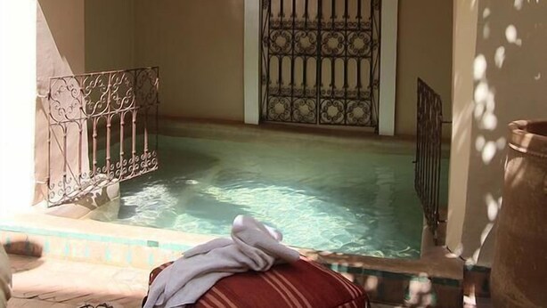 Un espace extérieur avec une piscine, un sol carrelé, un coussin rouge, une paire de sandales et un chapeau posé par terre.