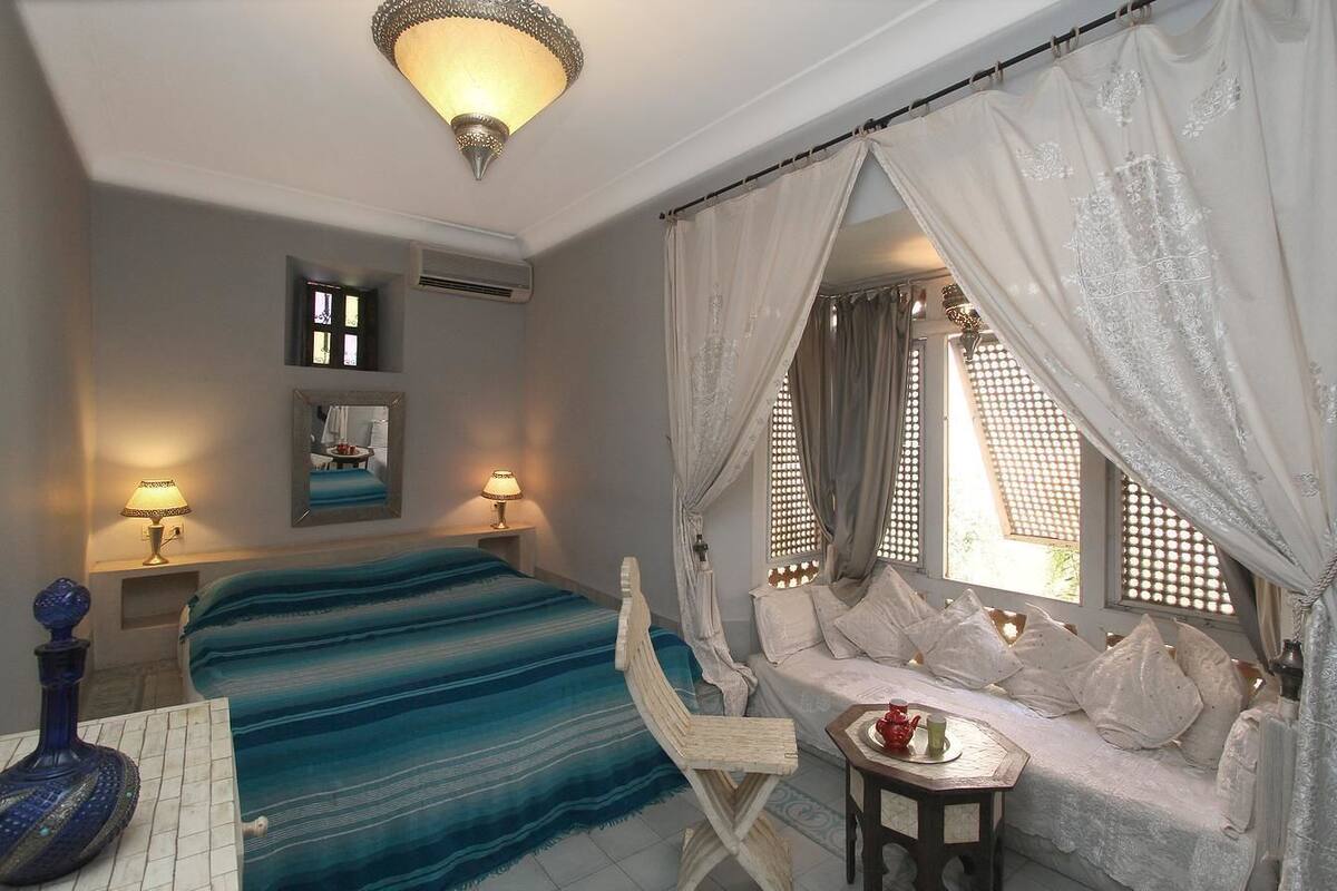 A bedroom with a bed, bedside lamps, a small table with a teapot, and a window with a view of the outside.
