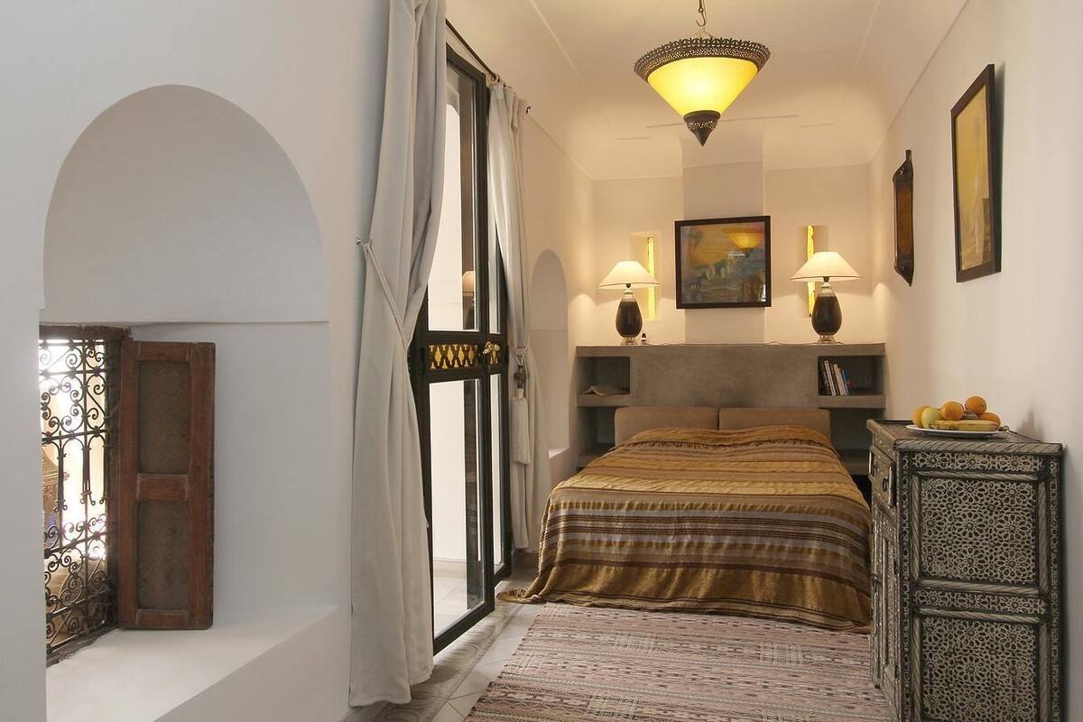 A bedroom with a bed, two bedside lamps, a nightstand, a decorative wall piece, and a view through a window with curtains.