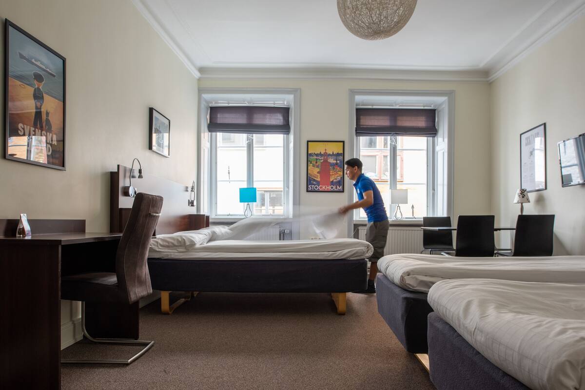 Habitación de hotel con dos camas, un escritorio y una silla. Dos ventanas con cortinas, un cartel en la pared y una lámpara sobre el escritorio.