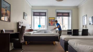 Ein Hotelzimmer mit zwei Betten, einem Schreibtisch und einem Stuhl. Es gibt zwei Fenster mit Vorhängen, ein Poster an der Wand und eine Lampe auf dem Schreibtisch.