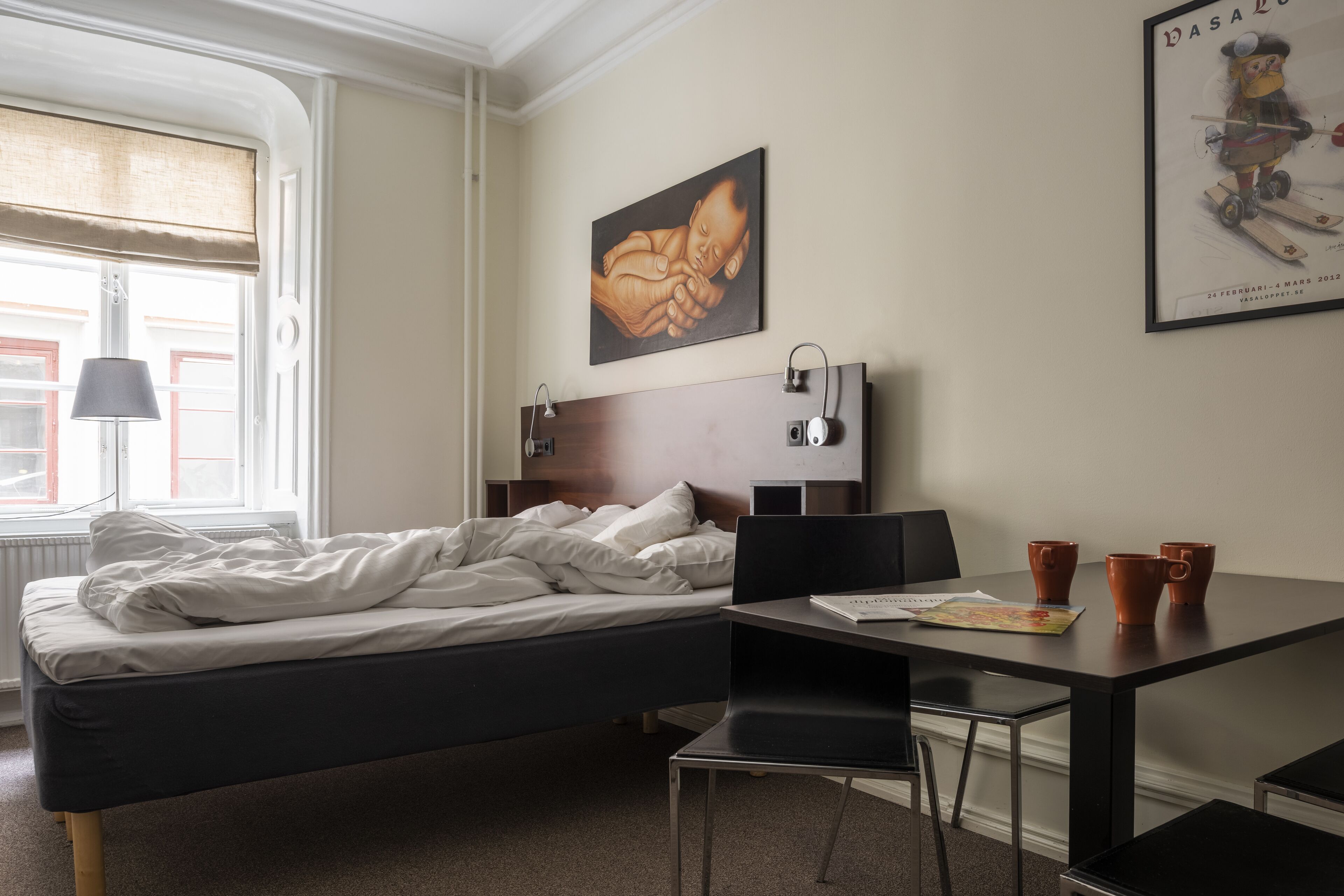 Habitación de hotel con una cama, un escritorio con dos sillas, una lámpara y un cartel en la pared.