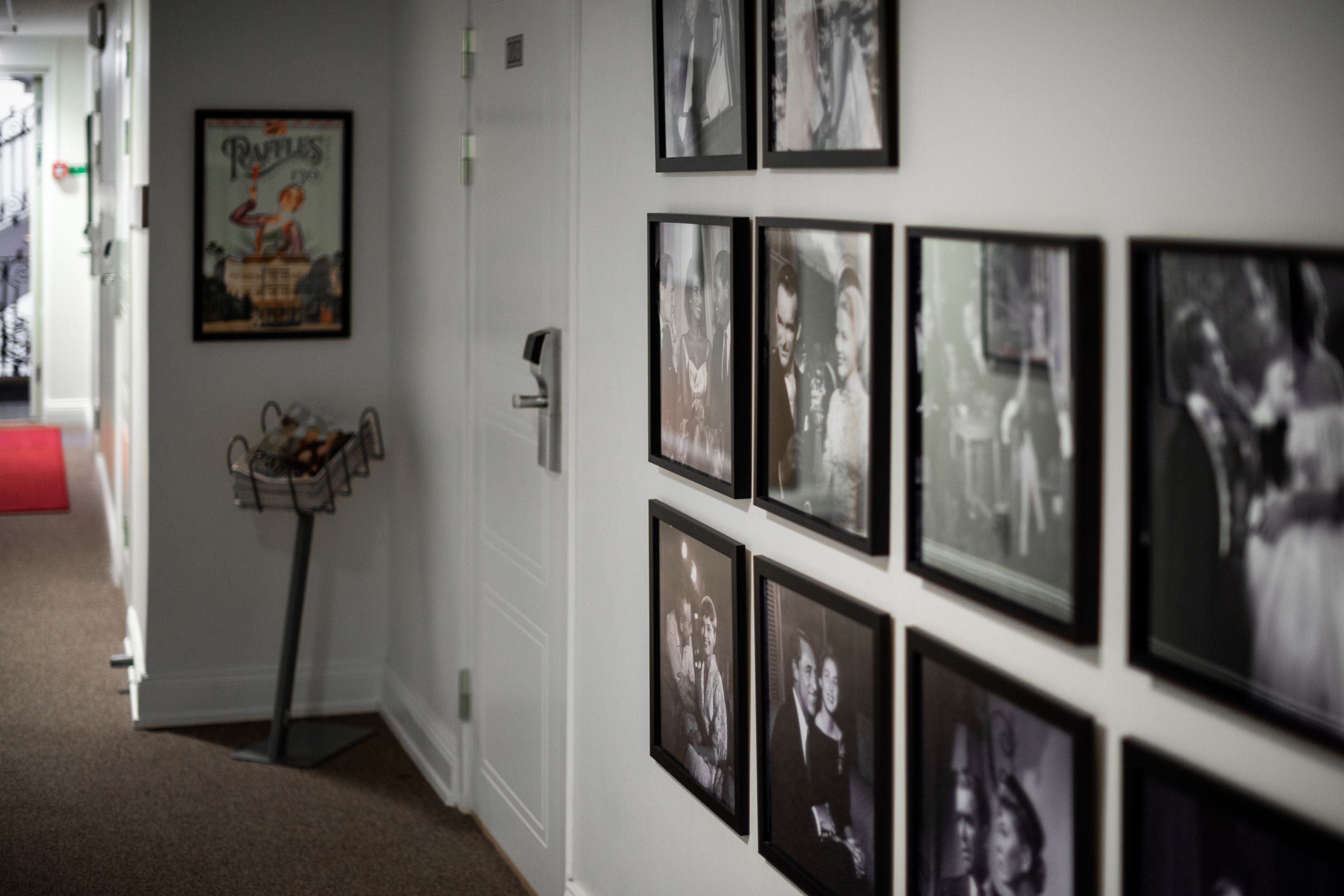 Un pasillo con fotografías blanco y negro enmarcadas en la pared, una pequeña mesa con una cesta y un objeto rojo en el suelo.