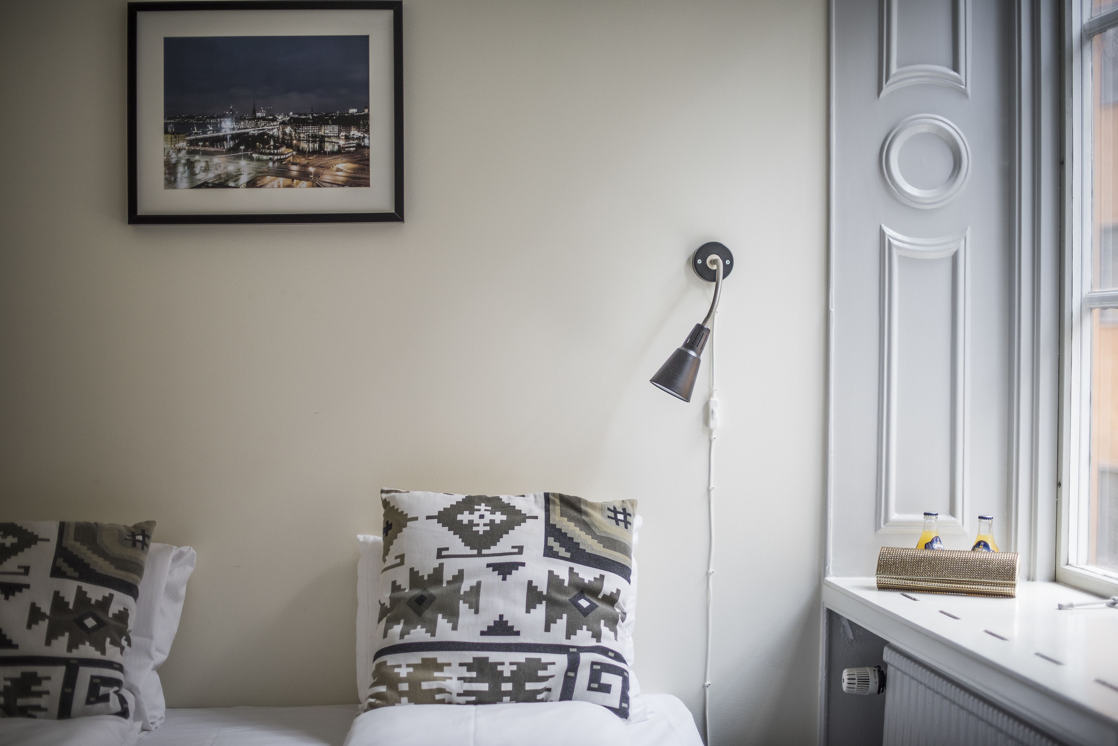 A neatly arranged bedroom with a bed, pillows, a framed picture, a lamp, and a window with bottles on the sill.