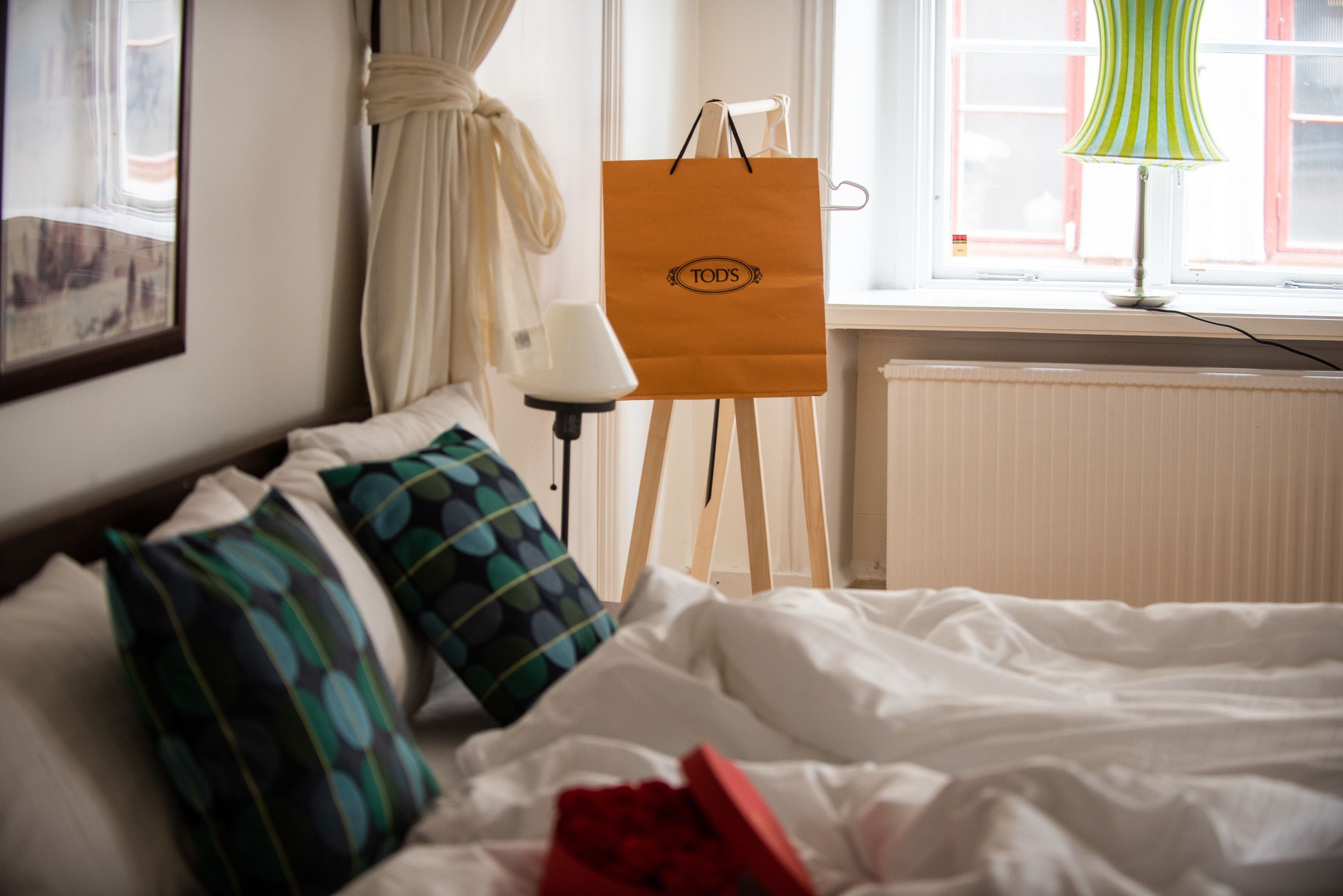 A hotel room with a bed, pillows, a standing lamp, a window with curtains, and a bag on a stand.