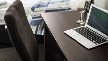 A laptop on a desk with a wine glass and a bottle, a bed with a striped blanket, and a window with a view of buildings.