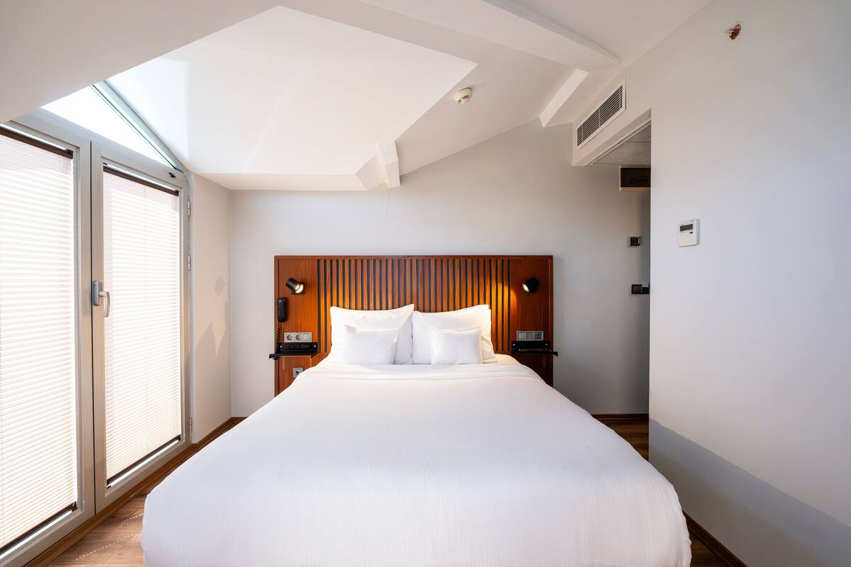 A modern hotel room with a large bed, skylight, wooden headboard, bedside lamps, and a sliding glass door.