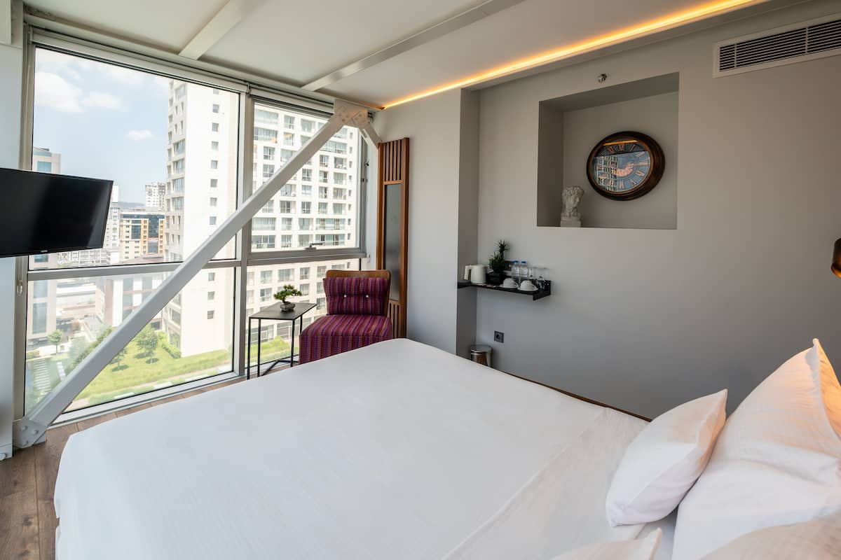 A modern hotel room with a large bed, a TV, a city view, a small table, and a chair.