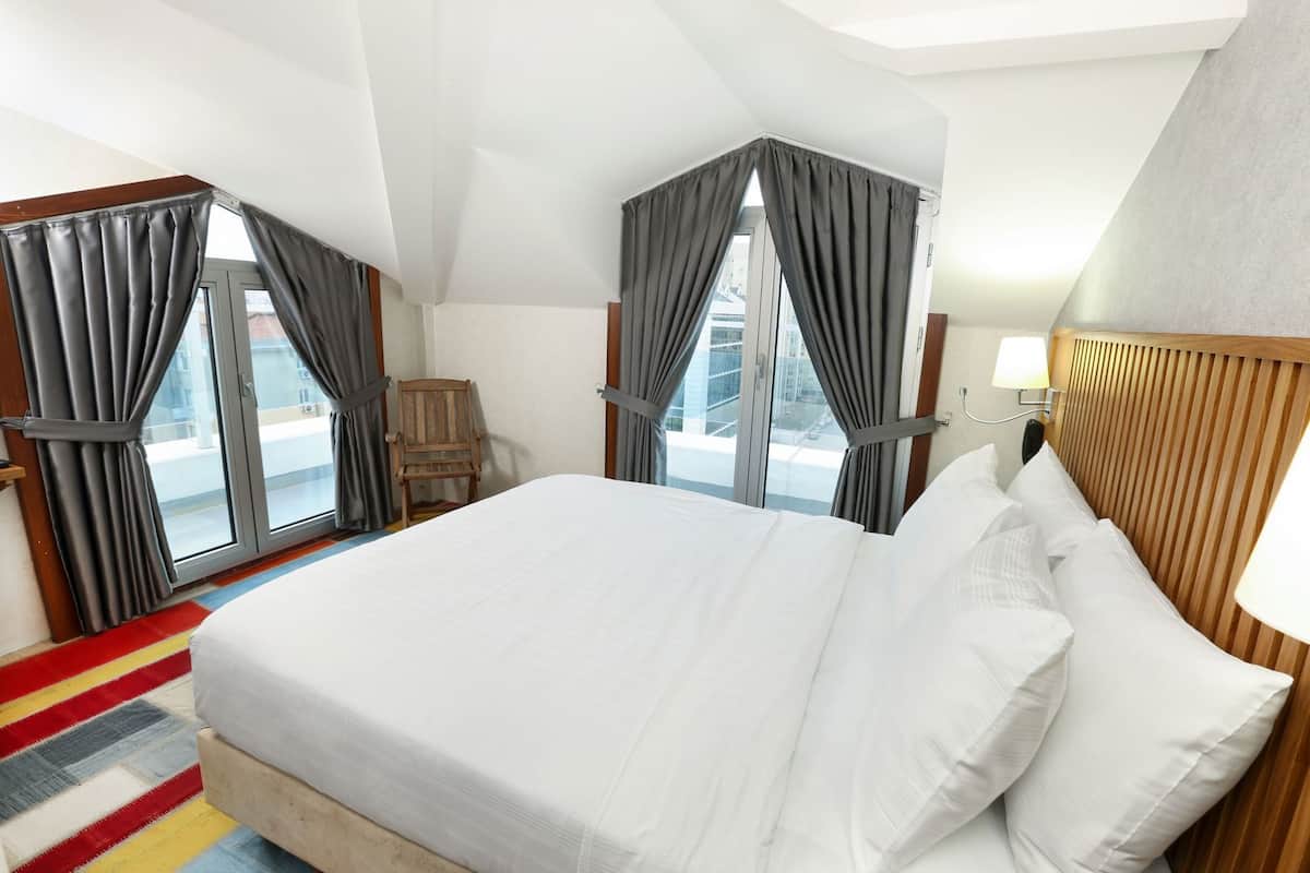 A hotel room with a large bed, two windows with curtains, a wooden headboard, a chair, and a colorful rug.