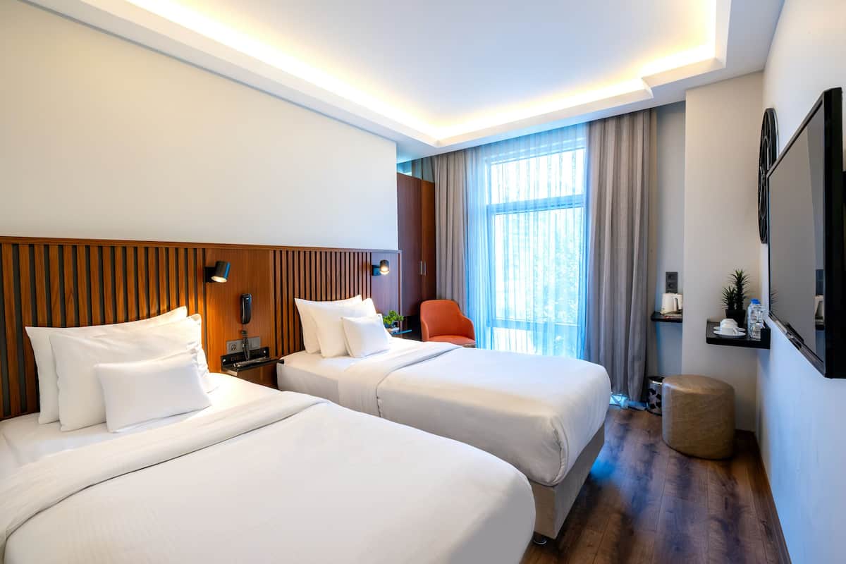 A hotel room with two beds, a wooden headboard, a nightstand, a chair, a round ottoman, a TV, and a window with curtains.