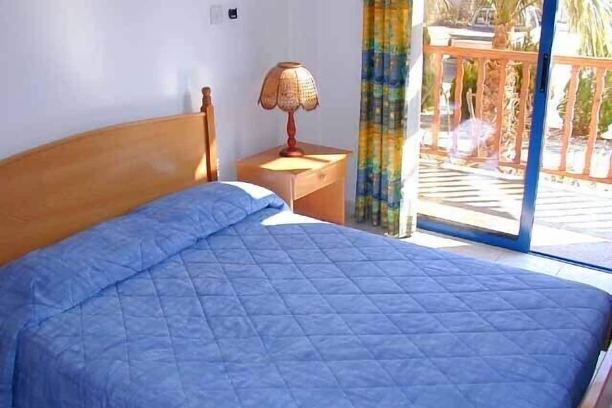A bed with a blue quilt, a wooden headboard, a nightstand with a lamp, and a view of an outdoor balcony with curtains.