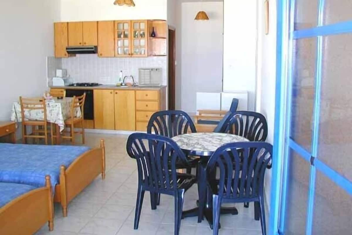 A compact room with a bed, a dining table with chairs, a kitchenette, and a small window.