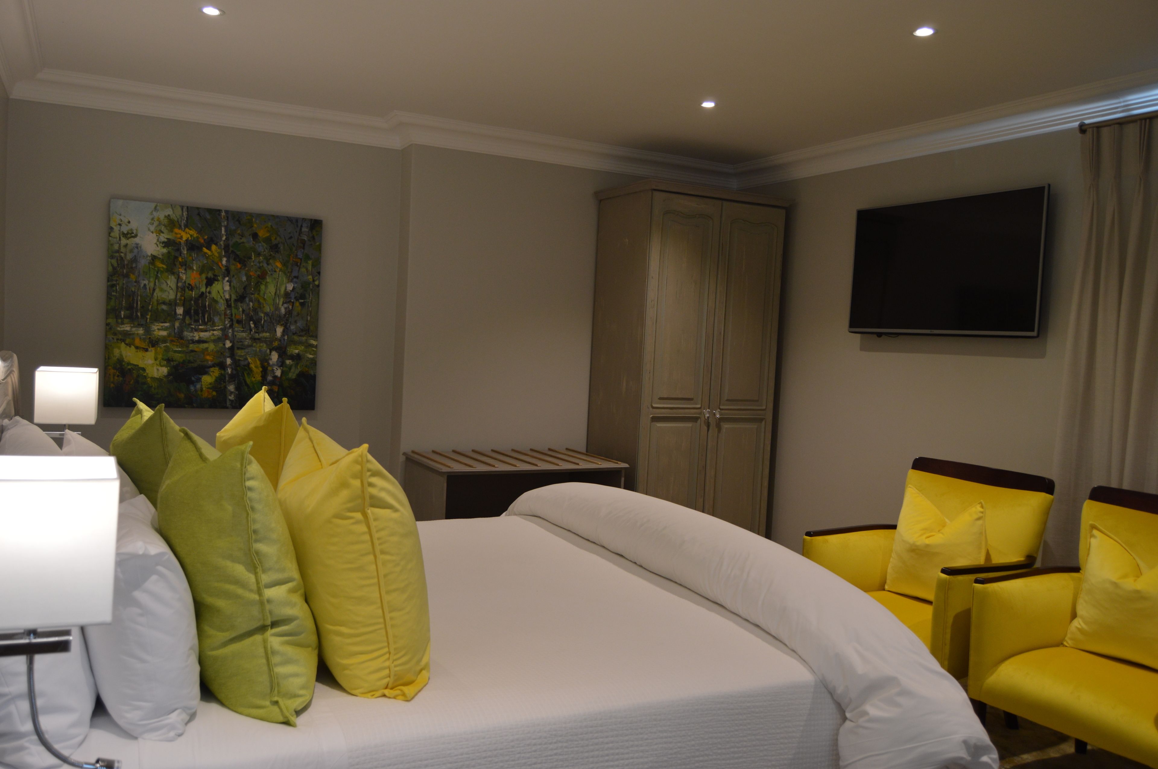 Une chambre d’hôtel avec un lit, des fauteuils jaunes, une télévision, un tableau et une armoire.