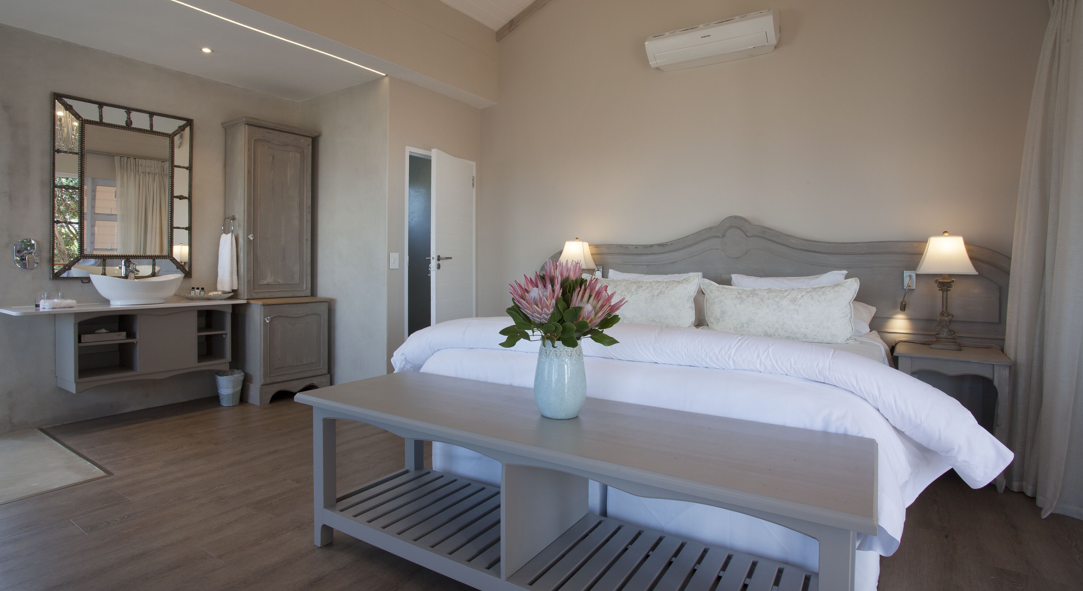 A hotel room with a bed, bedside lamps, a nightstand, a vanity with a mirror, a sink, and a vase with flowers.