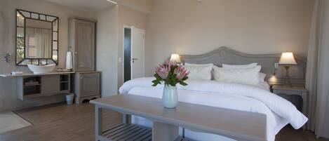 A hotel room with a bed, bedside lamps, a nightstand, a vanity with a mirror, a sink, and a vase with flowers.