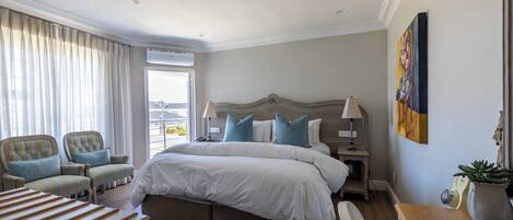 A hotel room with a large bed, two armchairs, a bedside table with a lamp, a painting on the wall, and a potted plant.