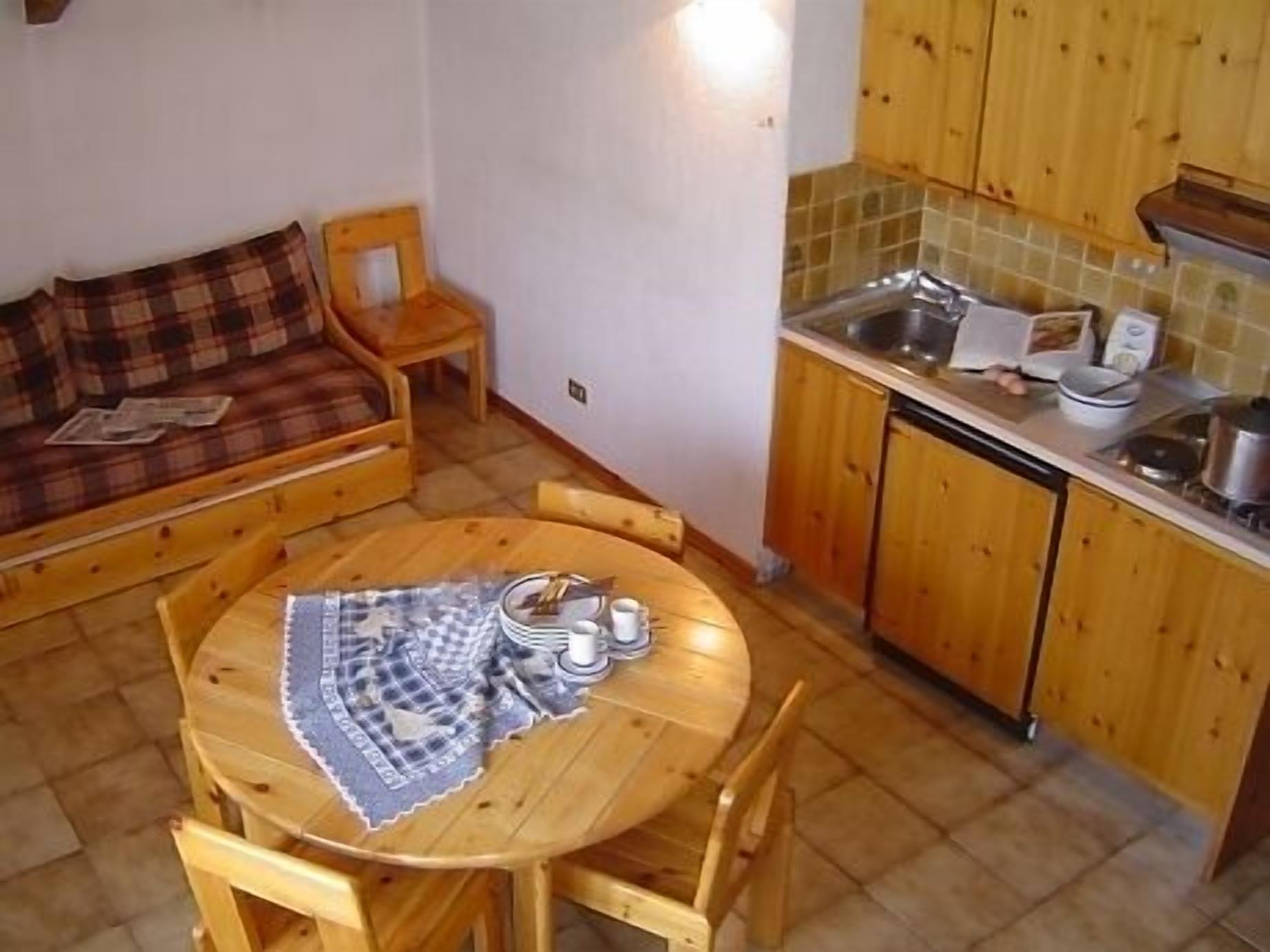 A cozy room with a wooden dining table set for two, a wooden sofa with a plaid blanket, and a kitchen area with wooden cabinets and a tiled backsplash.
