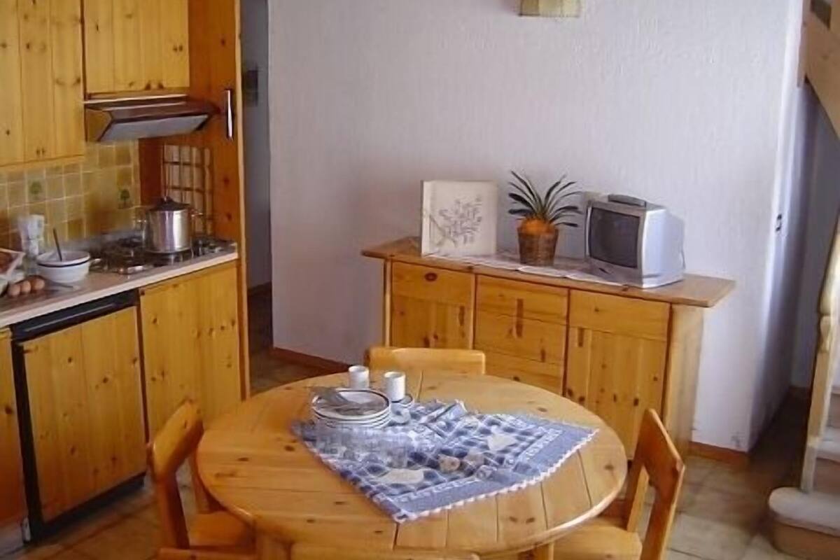 A small kitchen with wooden cabinets, a dining table with chairs, a microwave, and a potted plant.