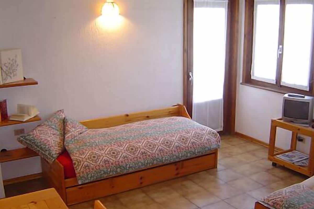 A small, cozy room with a single bed, a wooden table, a television on a stand, and a wall-mounted light.