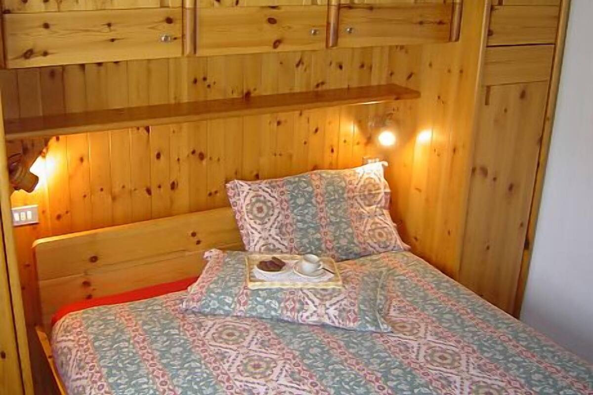 A wooden cabin room with a bed, a shelf above the bed, and a wall with wooden cabinets.