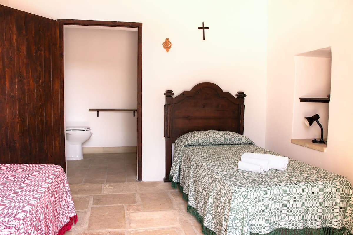 Quarto com cama de cabeceira de madeira, mesa de cabeceira com abajur, colcha estampada em vermelho e branco, e colcha estampada em verde.