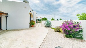 Un patio con un edificio blanco, un sendero curvo y un pequeño jardín con flores moradas.
