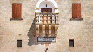 Fachada de edificio con una ventana central arqueada, flanqueada por dos ventanas más pequeñas con contraventanas de madera. Un balcón con barandilla decorativa está adosado a la ventana arqueada.