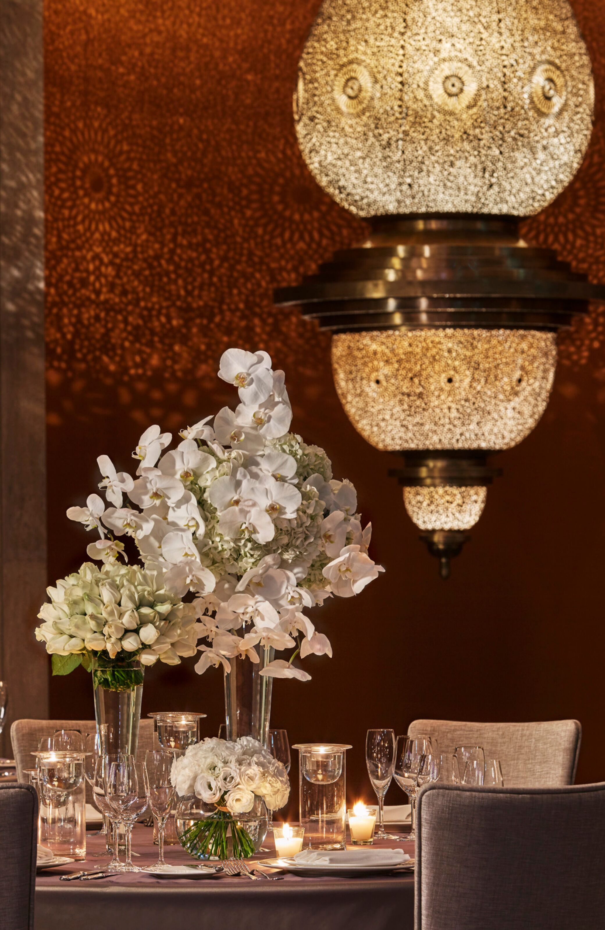 A dining table set with white flowers, clear glassware, and candles, with a textured wall and a spherical light fixture above.