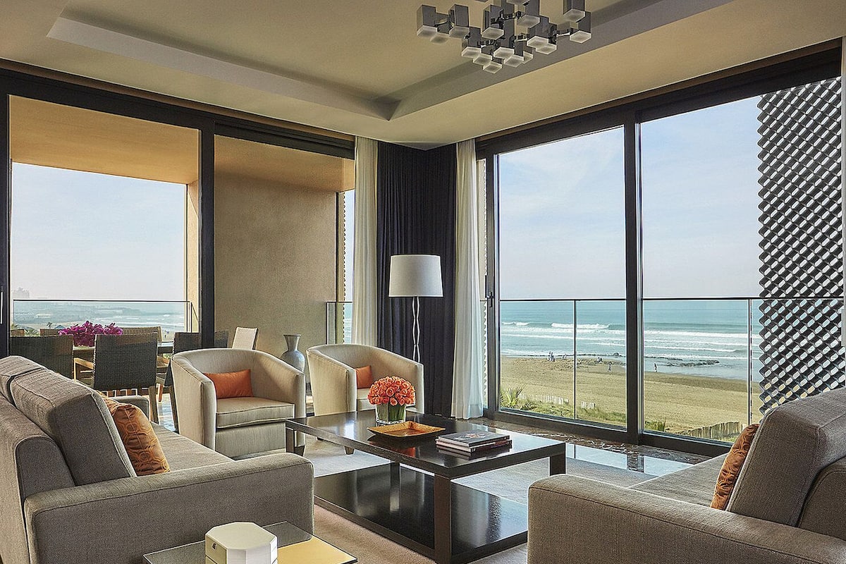 A modern living room with a view of the beach, featuring a sofa, armchairs, a coffee table, and a lamp.