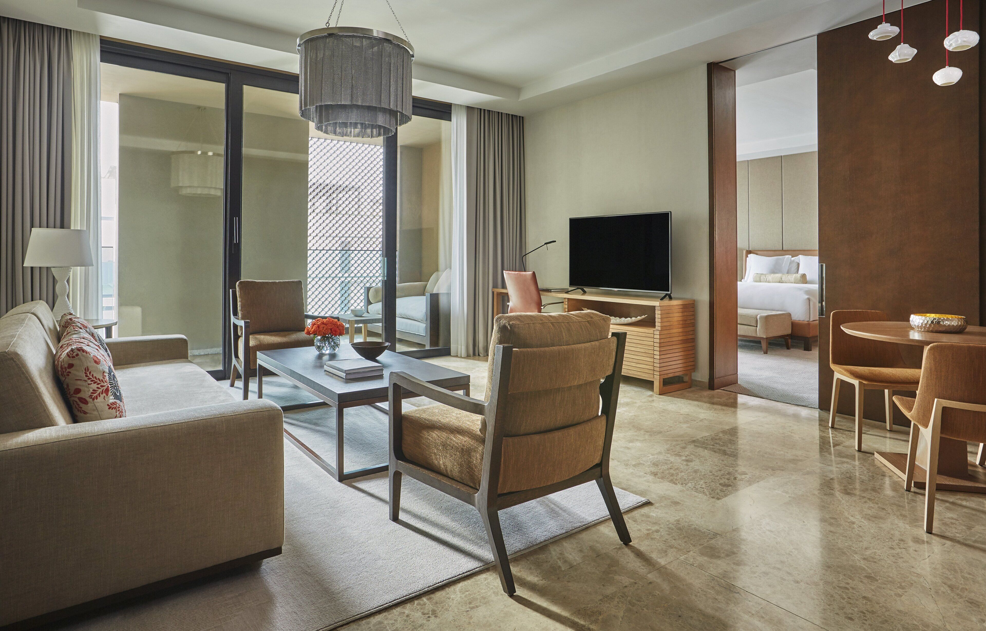 A modern hotel room with a sofa, armchairs, a coffee table, a TV, and a dining area with a table and chairs.