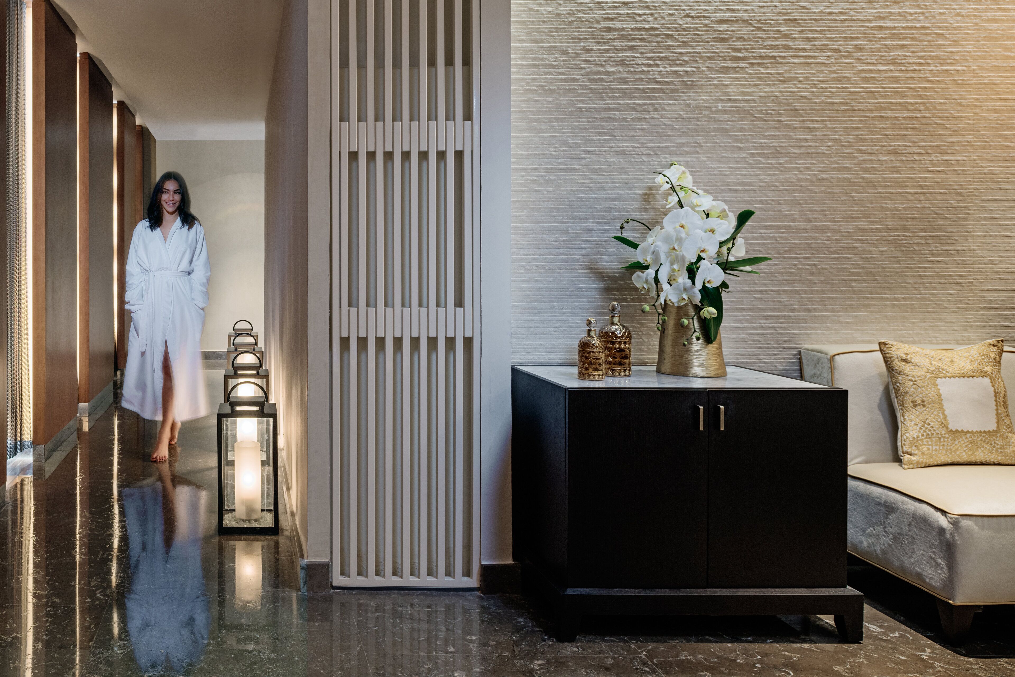 A hotel corridor with a reflective floor, a white robe-wearing person, a black cabinet with decorative items, and a sofa with a patterned cushion.