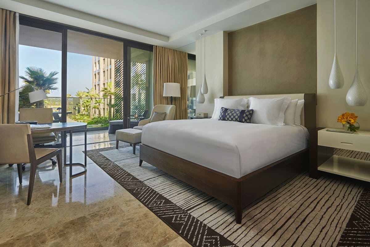 A hotel room with a large bed, a desk, and a chair. There is a view of palm trees and buildings outside the window.